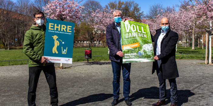 Drei Männen stehen nebeneinander in einer Parkanlage und halten Plakate, die für mehr Sauberkeit in Parks werben