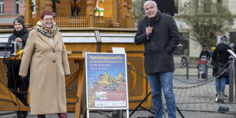 Eine Frau und ein Mann stehen links und rechts von einem Plakat in belebtem Umfeld. 