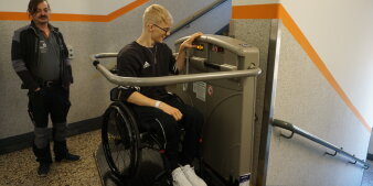 Ein Rollstuhlfahrer testet einen neuen Fahrstuhl. Daneben steht ein Mann in Arbeitskleidung. 