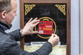 ein Mann bringt einen Aufkleber mit dem Schriftzug "nette Toilette" an einem Glaskasten an