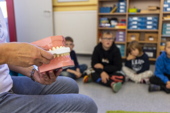 Eine Frau erklärt an einem Modell das Zähneputzen. Im Hintergrund sitzen ein paar Kinder und hören zu.