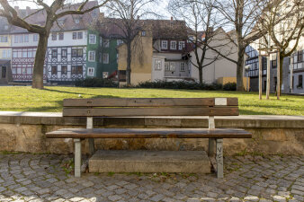 Eine leere Bank, im Hintergrund Bäume, Wiese und Häuser