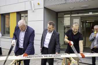 Zwei Männer sägen gleichzeitig an einem Brett. In der Mitte steht ein dritter Mann und beobachtet dies. 