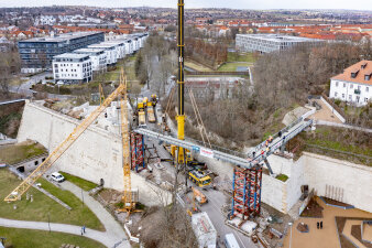 Luftaufnahme einer Baustelle, an einem Kran hängt ein Stahlteil für eine Brücke