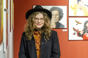 Eine junge Frau mit lockigen Haaren und Hut steht lächelnd vor gerahmten Zeichnungen 