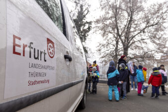 Zwei Frauen und zahlreiche Kinder packen Geschenke in einem Kofferraum. Im Vordergrund ein Auto mit Aufschrift. 