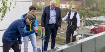 Vier Männer bei einer Richtfestzeremonie. Einer davon schwingt einen Hammer, um damit einen Nagel zu versenken.