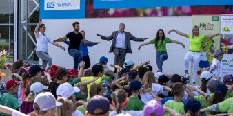 Mehrere Personen turnen auf einer Bühne. Davor stehen viele Kinder.