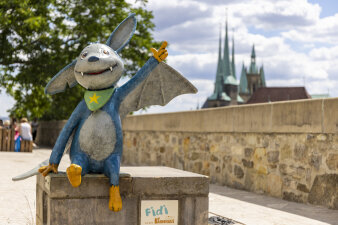 eine Fledermaus-Figur sitzt auf einem Podest, im Hintergrund der Erfurter Dom