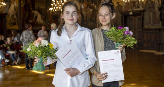 Zwei junge Frauen stehen mit Blumenstrauß und Urkunde beisammen.