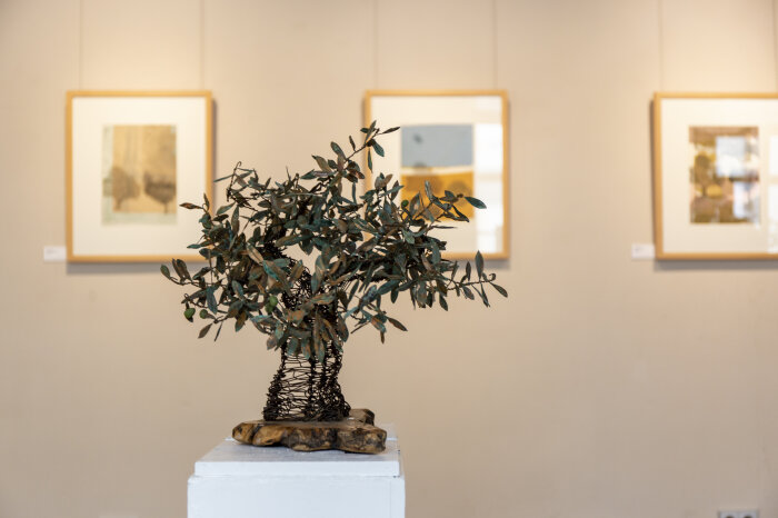 kleine Draht-Skulptur eines Olivenbaums, im Hintergrund hängen Fotos an der Wand