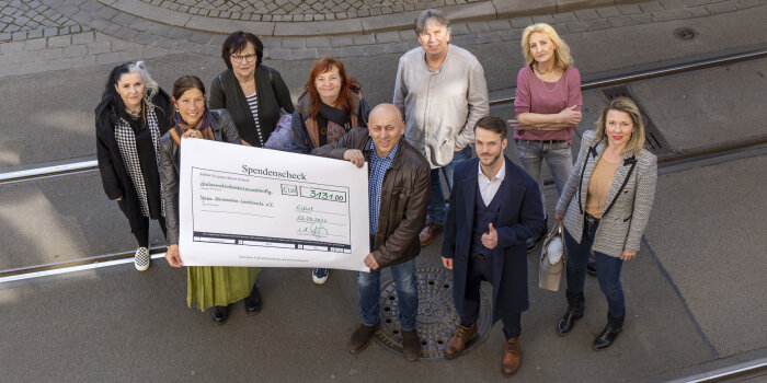 ein Gruppenfoto mit neun Menschen, zwei von Ihnen halten ein großes Papier, auf dem "Spendenscheck" steht
