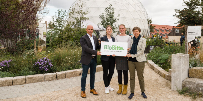 vier Personen stehen vor einem kuppelförmigen Bau und halten ein Schild mit Aufschrift "BioBitte" in der Hand