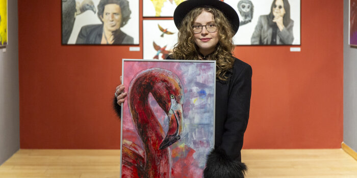 eine junge Frau mit Mantel steht in einem Ausstellungsraum und hält ein Bild in den Händen