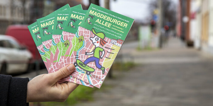eine Hand hält fünf Exemplare eines Faltplans mit Stadtkarte, Illustration und Titel "Magdeburger Allee"
