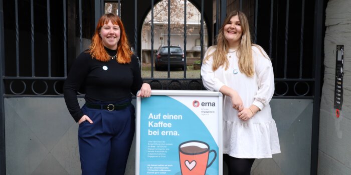 Zwei Frauen stehen an einem Aufsteller mit Plakat, auf dem "Auf einen Kaffee bei erna" steht.