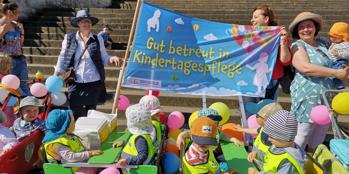 Vier Frauen, wovon zwei ein Plakat halten. Davor sitzen etwa 15 Kinder vor einer großen Treppe. 