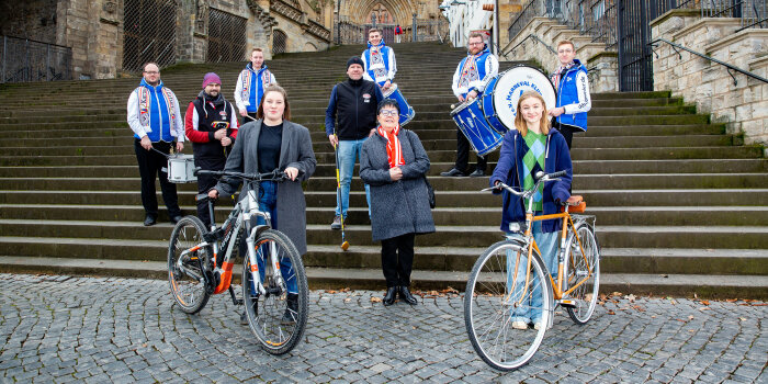 11 Menschen, darunter zwei Frauen mit Rad und Karnevalisten mit Trommeln, stehen auf und vor den Erfurter Domstufen
