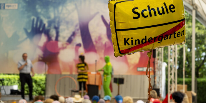 Viele Kinder sitzen vor einer Bühne. Im Vordergrund ist ein Ballon mit Aufschrift zu sehen. 
