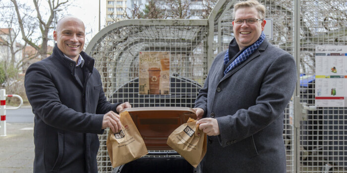 zwei Männer werfen jeweils eine Mülltüte aus Papier in eine Biotonne