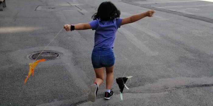 Ein Kind in Rückenansicht. Es probiert zwei gebastelte Flugdrachen auf dem Schulhof aus. 