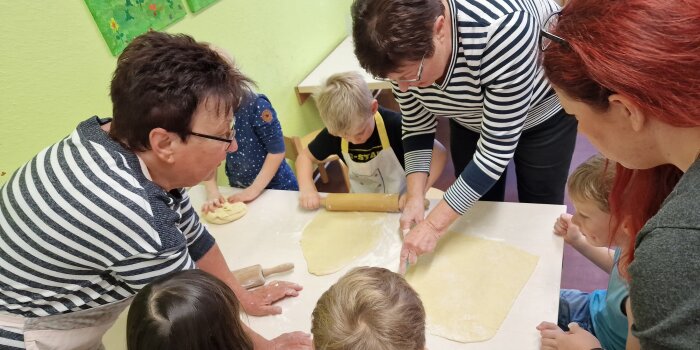 Drei Frauen und fünf Kinder stehen an einem Tisch und rollen gemeinsam Teig für Backwaren aus.