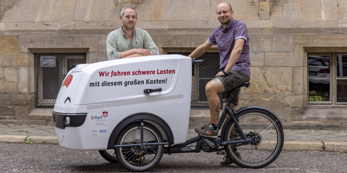 Ein Mann sitzt auf einem Fahrrad mit einem großen Transportkoffer, ein anderer Mann steht dahinter.