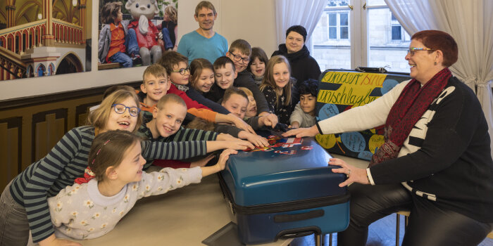 Eine Frau hat einen Koffer geöffnet und hält den Deckel fest. Ein paar Kinder schauen hinein.