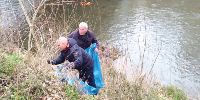 zwei Männer sammeln Müll in einer Böschung am Ufer eines Flusses