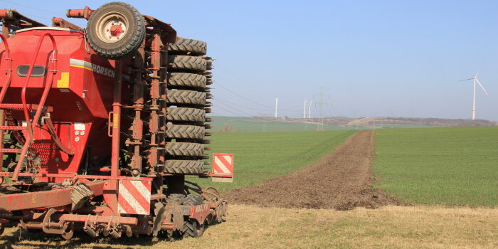 im Vordergrund eine große landwirtschaftliche Maschine, im Hintergrund ein Feld mit einem geackerten Streifen