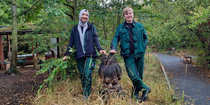 eine junge Frau und ein junger Mann stehen im Grünen neben einer Eulenfigur aus Holz