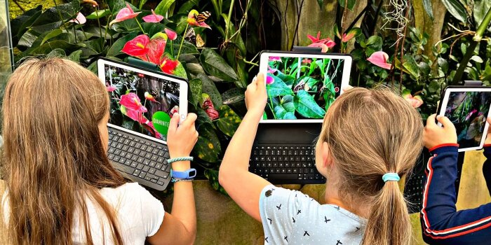 Zwei Mädchen stehen mit Tablets in der Hand vor einer großen Pflanze. 
