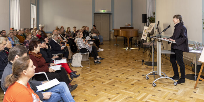 Eine Frau steht an einem Pult und spricht. Ihr gegenüber sitzen viele Menschen und hören zu. 