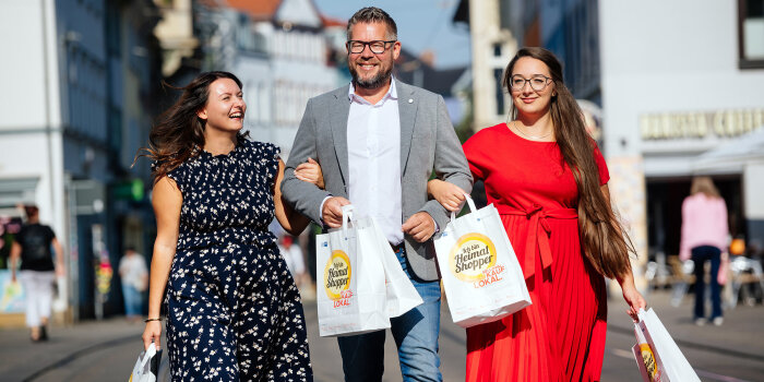 Foto: Heimat shoppen © Steve Bauerschmidt zwei Frauen haben sich bei einem Mann eingehakt und spazieren mit Papiertüten in der Hand