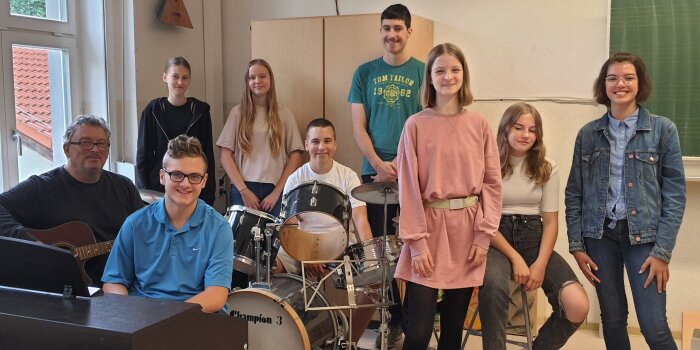 Acht jungen Menschen sitzen oder stehen zusammen mit ihrem Musiklehrer an beziehungsweise vor ihren Instrumenten.