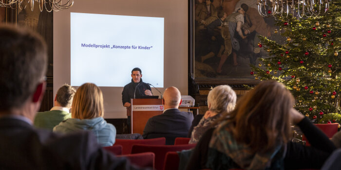 Ein Referent steht vor einer Leinwand in einem großen Saal. Ihm gegenüber sitzen viele Menschen auf Stühlen.
