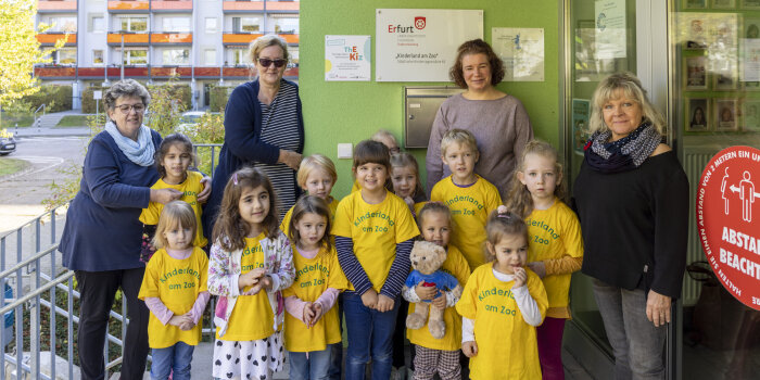 Eine Gruppe aus vier Frauen und zwölf Kindern steht vor einem Schild an einer Wand.