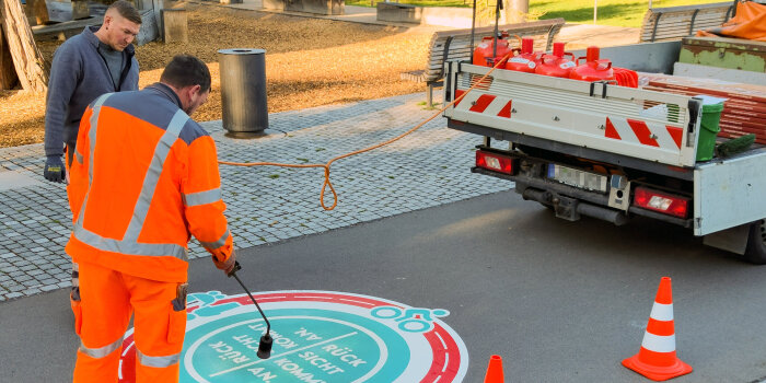 Zwei Personen bringen einen Aufkleber auf einer Straße auf.