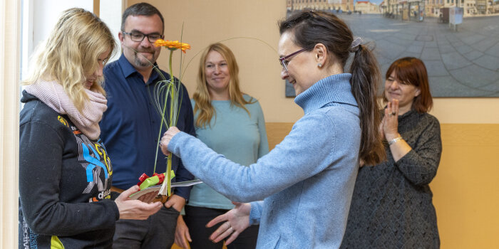 Eine junge Frau bekommt von einer Frau eine Blume überreicht. Im Hintergrund stehen ein Mann und zwei Frauen.