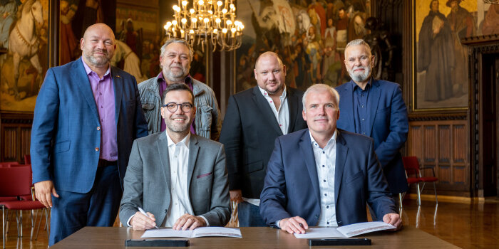 Zwei Männer sitzen an einem Tisch und halten jeweils einen Stift in der Hand. Dahinter stehen vier weitere Männer.