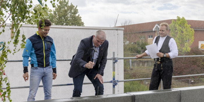 Drei Männer bei einer Richtfestzeremonie. Einer davon schwingt einen Hammer, um damit einen Nagel zu versenken.