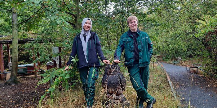 eine junge Frau und ein junger Mann stehen im Grünen neben einer Eulenfigur aus Holz