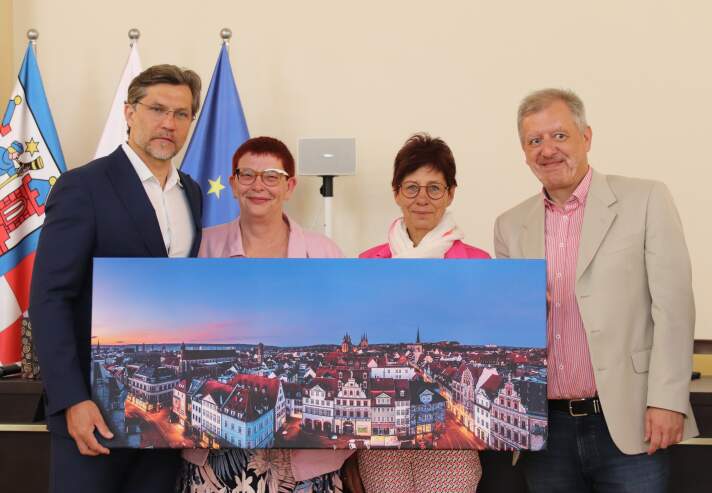 Zwei Männer und zwei Frauen halten ein Bild mit einer Stadtansicht. 