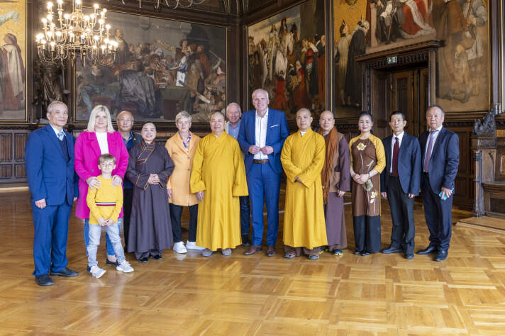 Ein Mann im Anzug sowie eine Frau und drei Männer in religiösen Roben sowie weiteren Besucher stehen beisammen.