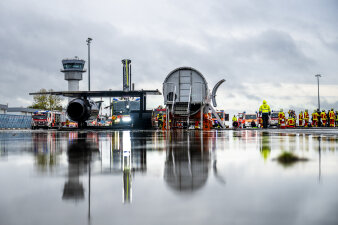 Auf einem Rollfeld stehen ein Flugzeugrumpf, Fahrzeuge und Menschen. Alles spiegelt sich auf dem feuchten Boden. 