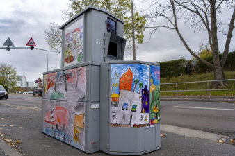 Ein Gerät zur Geschwindigkeitsmessung steht an einer Straße und ist mit bunten Bildern beklebt. 