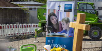 Ein Plakat einer Kirchenstiftung steht auf einer Baustelle, davor auch ein hölzernes Kreuz.
