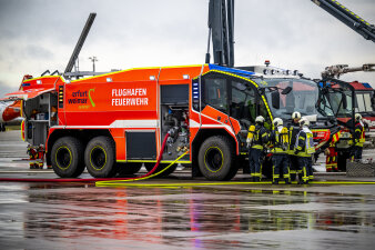 Ein Löschwagen der Flughafenfeuerwehr auf dem Rollfeld. Davor mehrere Personen. 