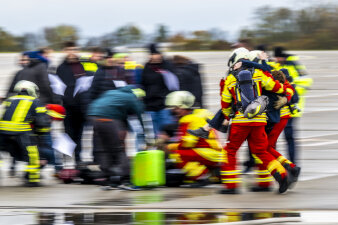 Eine verschwommene Aufnahme in Aktion. Mehrere Statisten und Feuerwehrleute auf einem Rollfeld. 