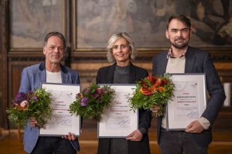 drei Personen halten je einen Blumenstrauß und eine gerahmte Urkunde